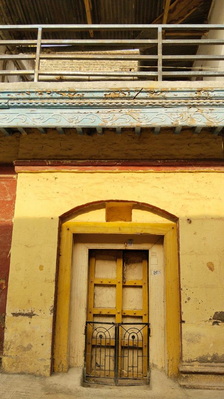 Main entrance with yellow-painted wooden panels and decorative elements. Above the door, a carved niche, distinct in color, is traditionally used to place an image or murti of a devi or devta with the belief that they will protect the home. A half-height railing is attached to the door as a safety barrier. (Source: CKA Archives)