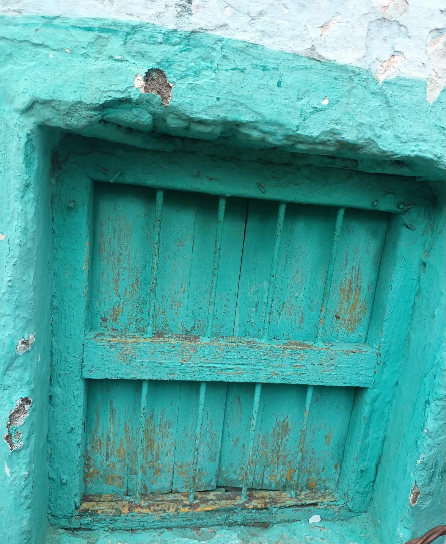 A horizontal ventilation window painted in the same turquoise color as the wall. It features a simple slatted wooden design, likely for air circulation. The window appears to be recessed into the wall and shows considerable aging. (Source: CKA Archives)
