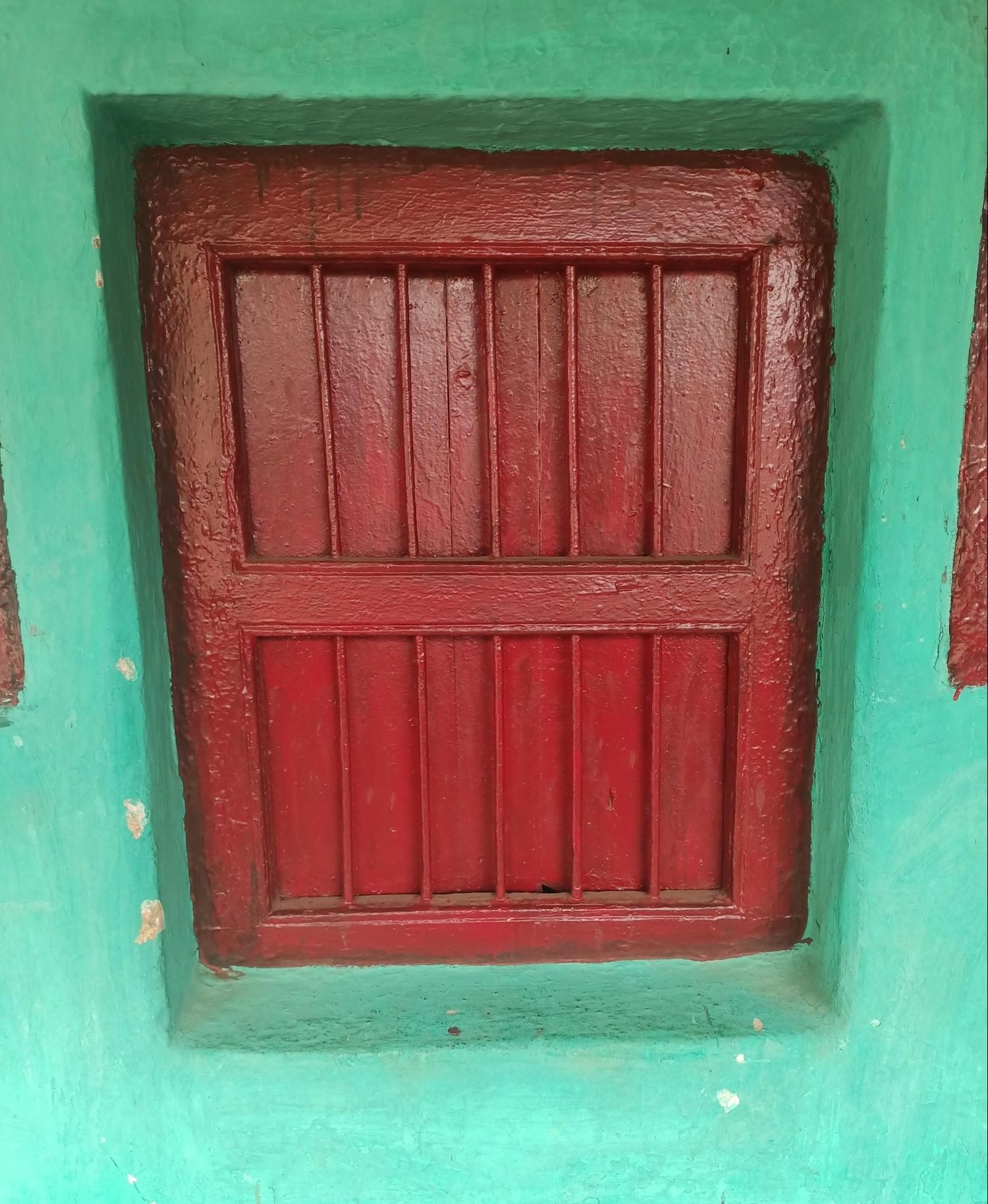 A red-painted wooden window with vertical planks divided into two sections (upper and lower panels), set within a green-painted wall frame. (Source: CKA Archives)