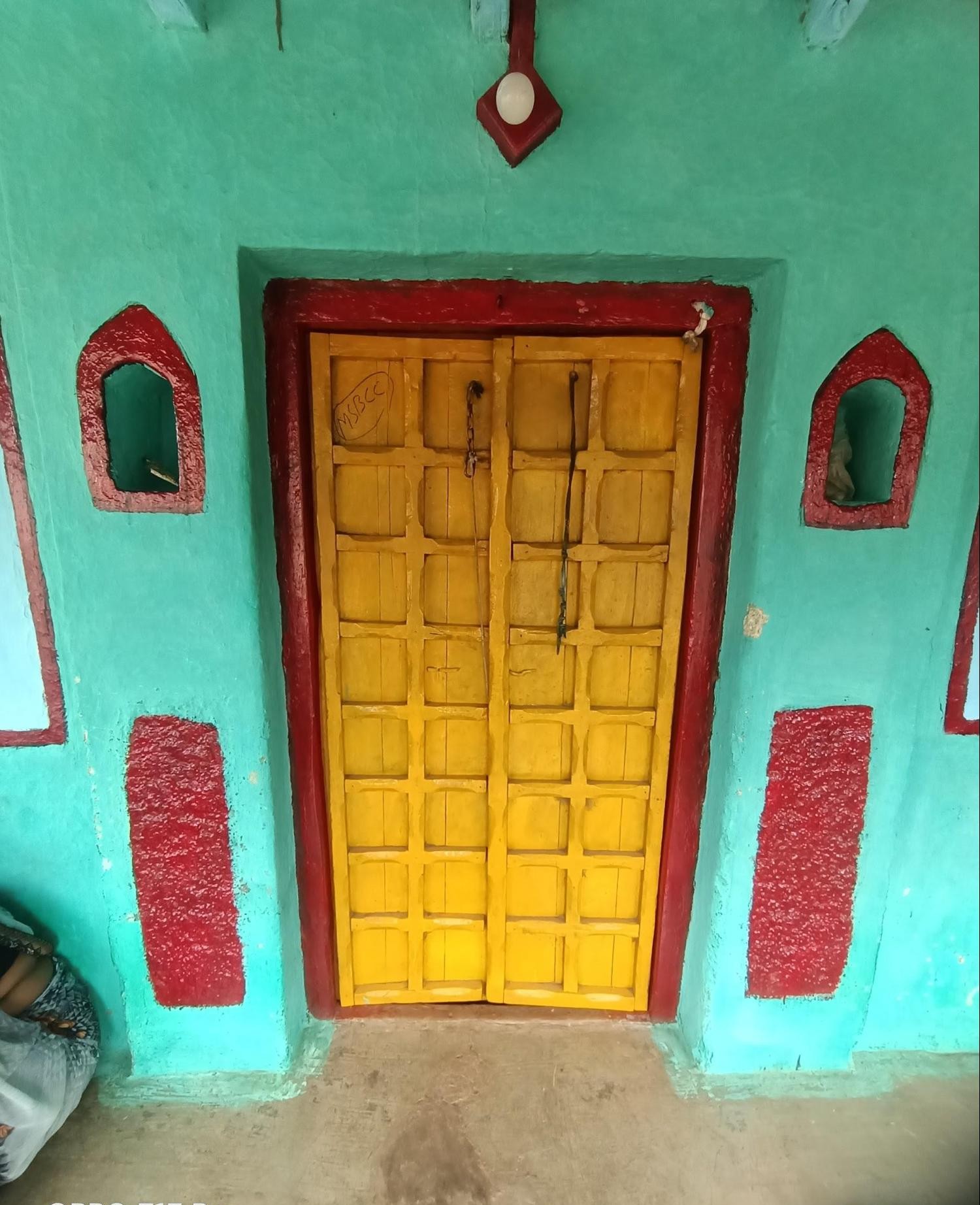 Yellow wooden door with paneled design, framed by red trim and flanked by decorative red-painted niches and rectangular panels on a turquoise wall. A pendant-style light fixture hangs above. (Source: CKA Archives)