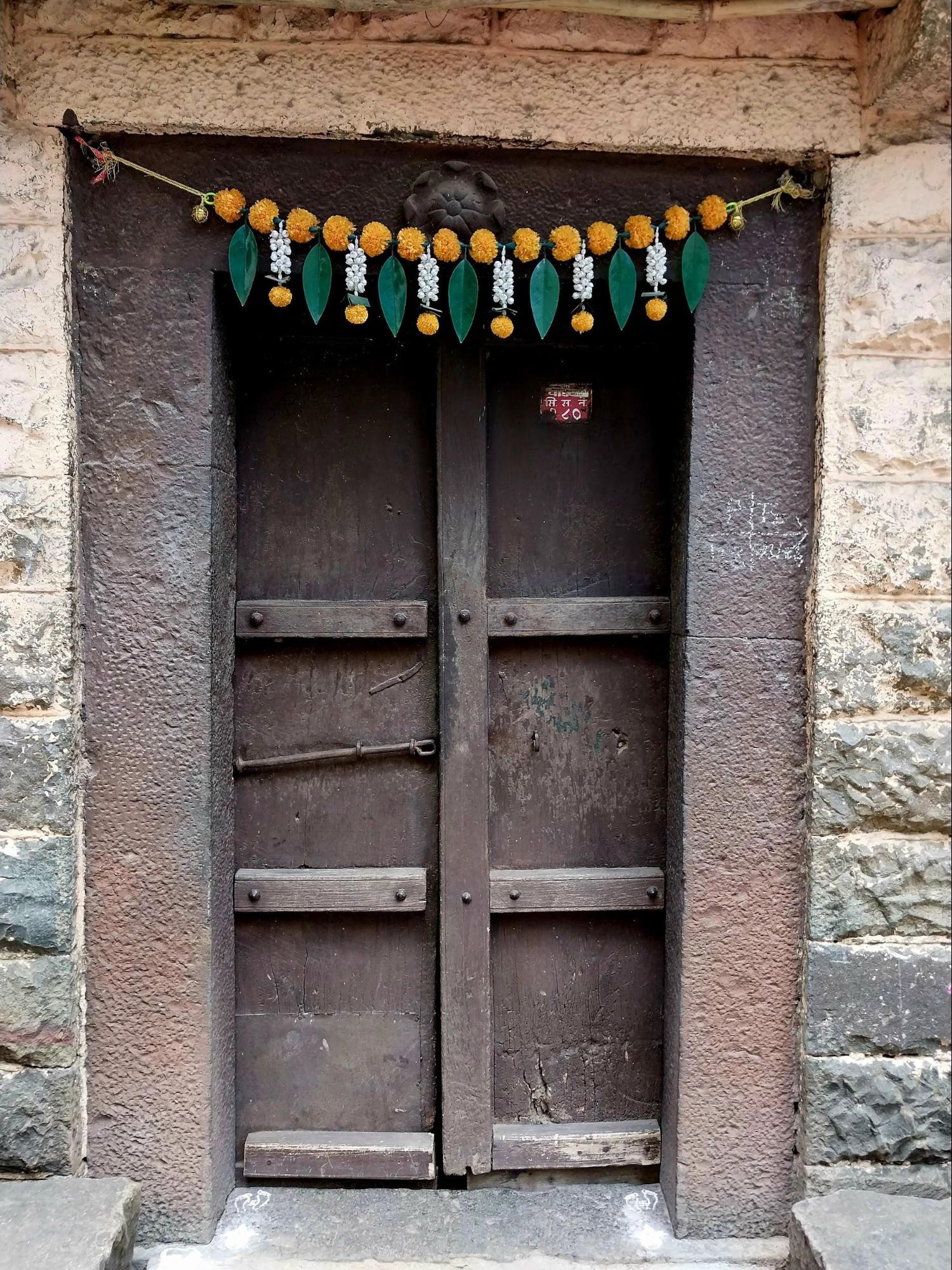 The entrance door of the residence is adorned with a toran, a traditional cultural element. Notice the stone walls on either side, as well as the construction of the door alongside details like wooden planks and visible screws. (Source: CKA Archives)
