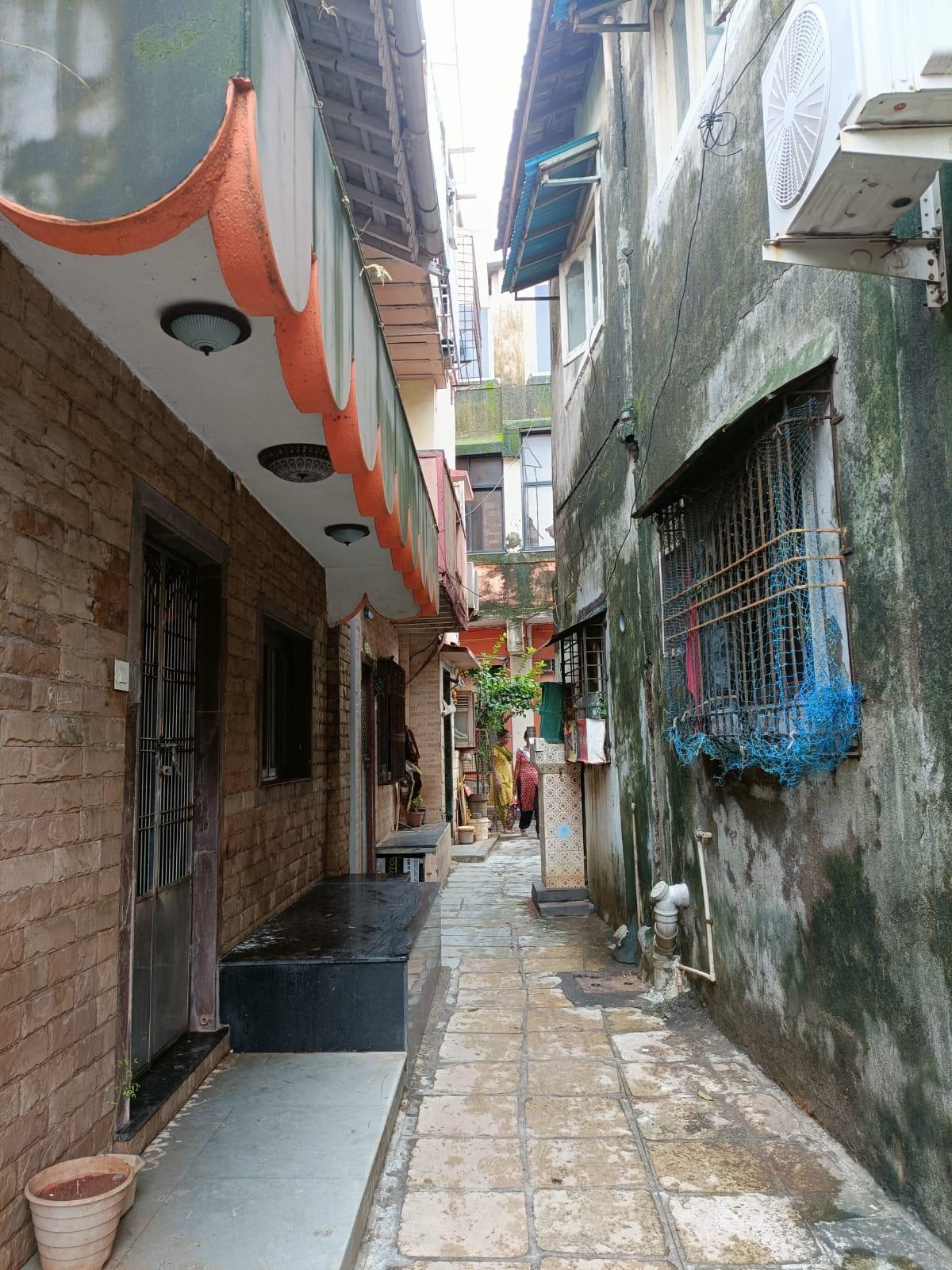 Residence (right) with exposed brickwork walls, a stone-tiled verandah, a metal door and a parapet with a vibrant orange design. Residence at (left) with an unfinished cement base. Windows on the bottom storey are enclosed with metal grills, while windows on the top storey have shutters that open outward. (Source: CKA Archives)