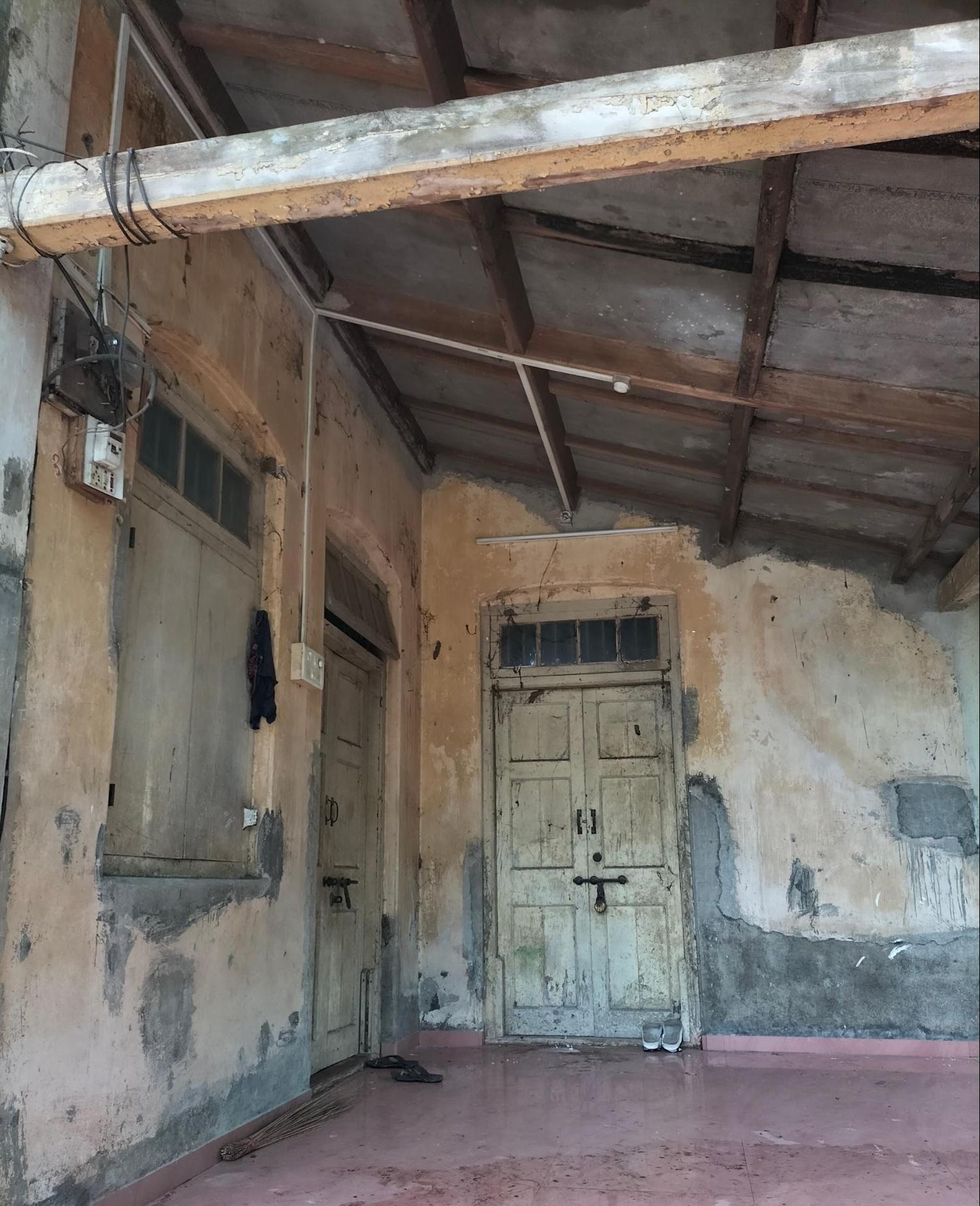Close-up of the entrance doors showing signs of age and everyday use. The wooden double-leaf doors are set within a plastered wall, with fading paint, weathered surfaces, and small repairs visible across the frame. (Source: CKA Archives)