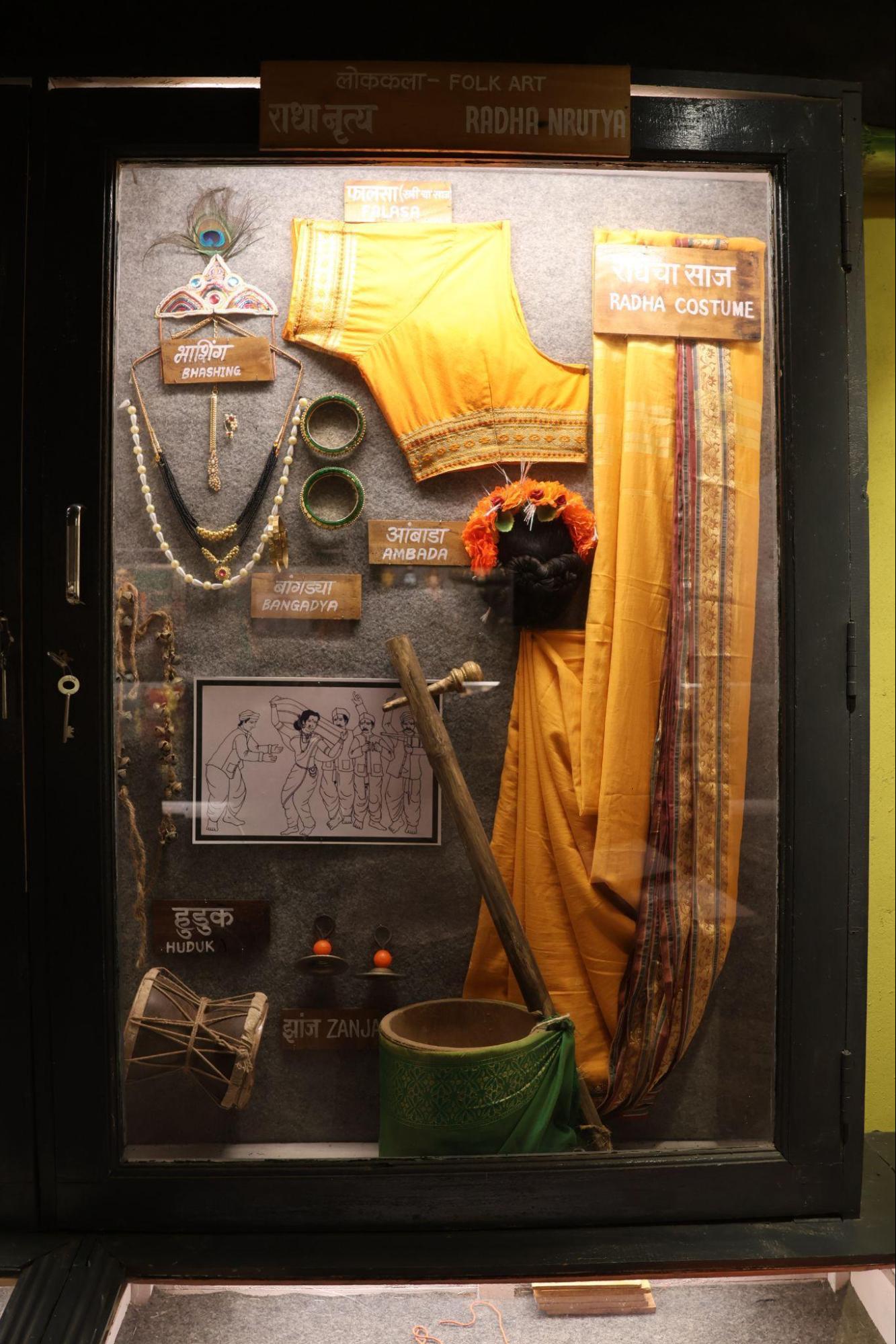 A display of costumes and instruments used in Radha Nritya, a folk dance performed in the Konkan region. Featured here are traditional jewellery pieces like bashing and bangadya, accessories like ambada (hair bun with flowers), and the vibrant Radha costume. Musical instruments such as the huduk drum and zanja (cymbals) accompany the performance, while a sketch illustrates a typical dance scene. (Source: CKA Archives)