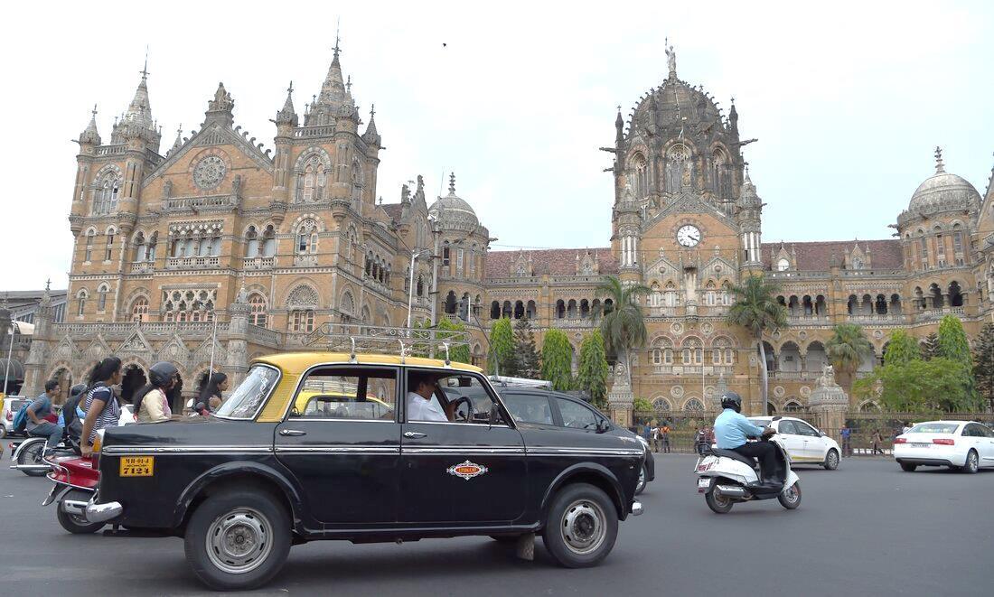 A Premier Padmini black-and-yellow taxi driving past the Chhatrapati Shivaji Maharaj Terminus, each an ever-present identity of Mumbai.